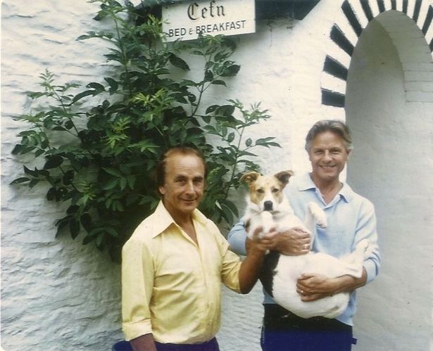 The image shows Reg and George in colourful clothing outside their B&B in Machynlleth