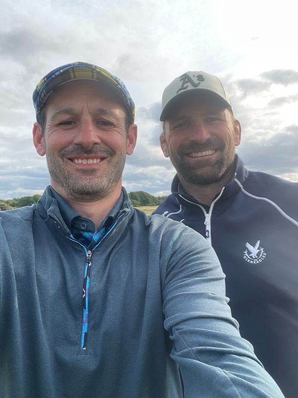 An image of two middle-aged men wearing zip-up jackets and caps, smiling into the camera.