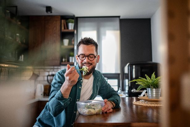 Young man with glasses and a beard is sitting at his kitchen counter, savoring a healthy salad from a plastic container, enjoying a mindful moment during his meal