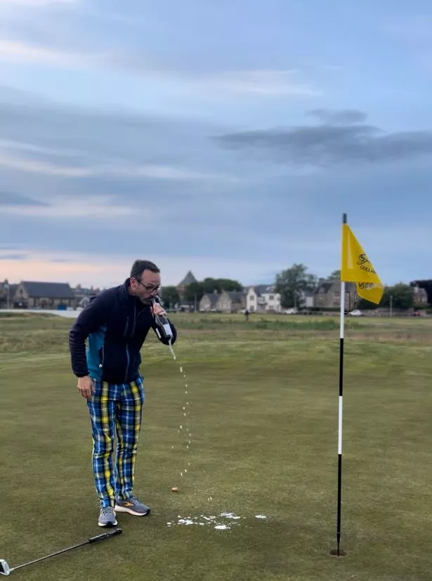 An image of a man wearing a blue fleece and blue and yellow tartan trousers, drinking from a bottle of champagne while standing on a golf course.