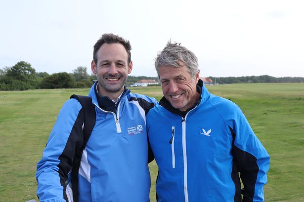 An image of a dark-haired man and older grey-haired man wearing blue waterproof jackets, standing on a golf course.