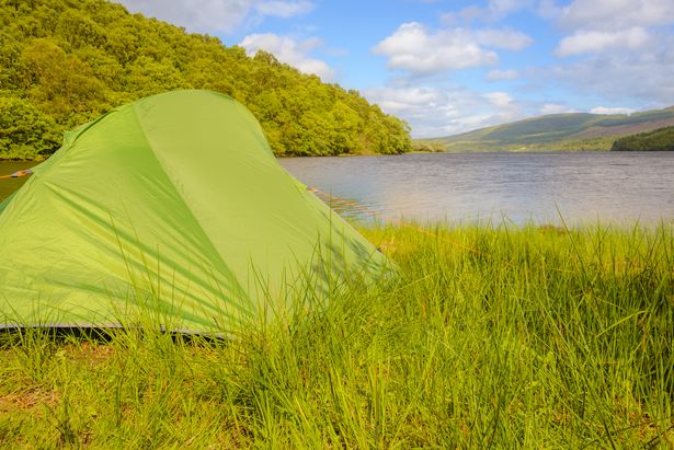 A tent pitched beside a summer scene in the Scottish Highlands, sun hits the forest-covered mountains and a large Loch is in the foreground.