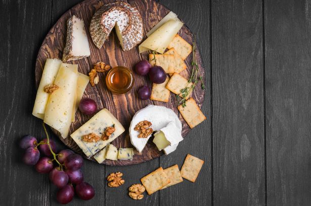 Assorted cheeses on round wooden board plate Camembert cheese, grated bark of oak, hard cheese slices, walnuts, grapes, crackers, bread, thyme, dark black wood background, top view