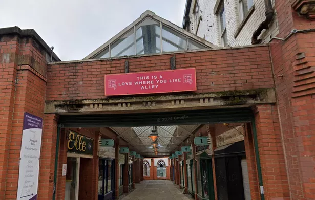 The shopping arcade behind the Manchester Chambers with a sign that reads 'This is a Love where you Live alley' hanging over a deteriorated concrete beam. 