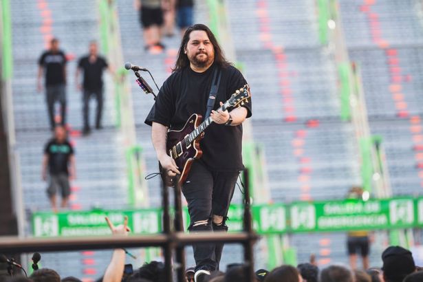 Wolfgang Van Halen in a black top and jeans on stage with a guitar.