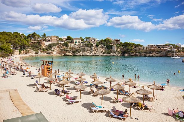 Vacationers relaxing under umbrellas and enjoying calm, crystal clear water on a sandy beach. Homes on cliffs in background.
