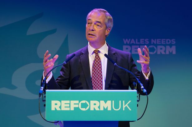 A man in a suit standing at a turquoise stand and speaking into two microphones
