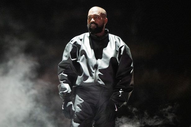 Kanye West in a black jacket and trousers, with gloves, on stage in 2024.
