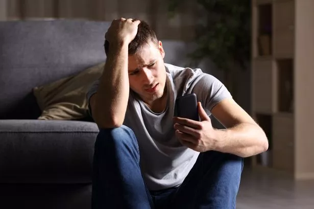 A depressed and anxious young man sat on the living room floor holding his phone