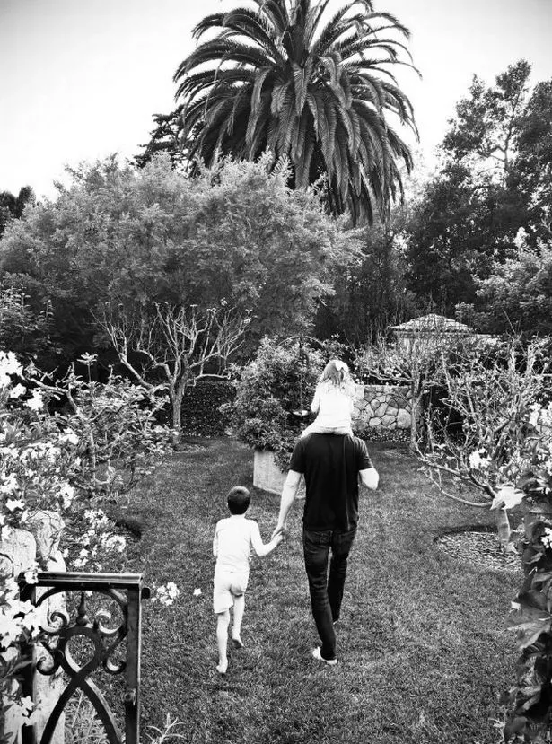 Black and white photo of Harry carrying Lilibet on his shoulders and holding Archie's hand in a garden, backs to the camera.