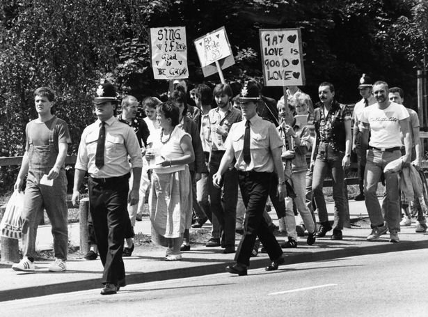 The image shows protesters walking with placards and surrounded by police