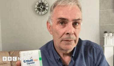 Paul Elcombe pictured in his living room holding up a box of Creon tablets. He is wearing a blue spotted shirt.