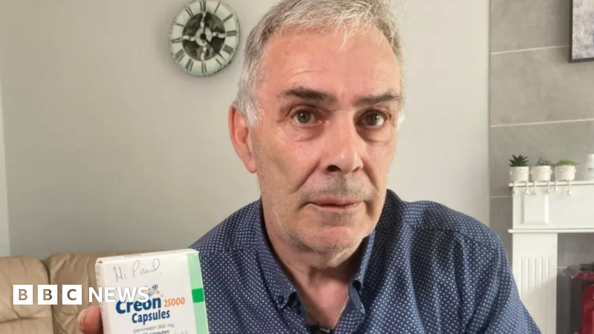 Paul Elcombe pictured in his living room holding up a box of Creon tablets. He is wearing a blue spotted shirt.
