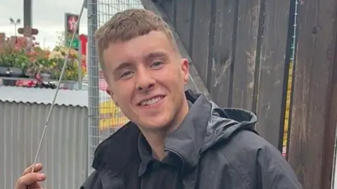 Tom McGrath Matthew McGrath has short brown hair, he is wearing a black polo shirt and black raincoat. Behind him is a brown wooden fence, a corrugated iron structure and a petrol station forecourt. 