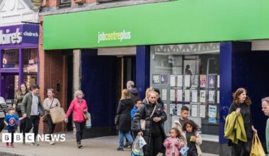 People, including families, walking on a street past the Job Centre Plus employment office in Chester. The street is busy, the weather is dry but people are wearing coats.