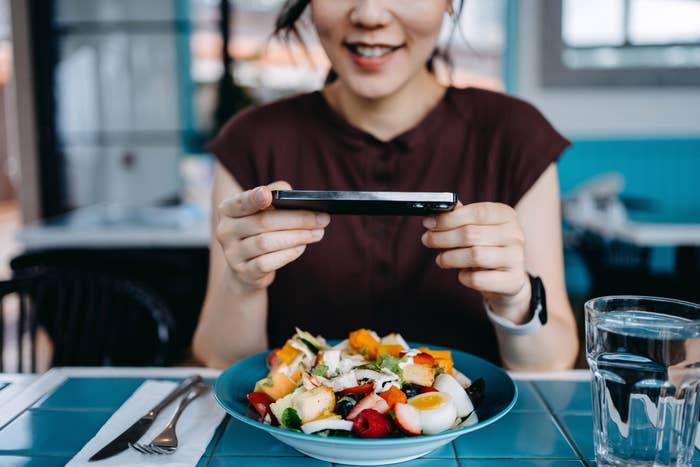 Person smiling while taking a photo of a colorful salad at a restaurant table with a smartphone