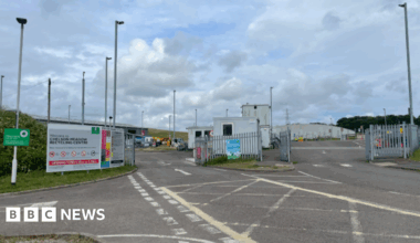 Entrance to Chelson Meadow Recycling centre. There is an entrance and exit gate for vehicles. A large sign says Welcome to Chelson Meadow Recycling Centre. There are several lampposts inside the site.