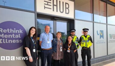 Three women and one man stand outside a building alongside two male police officers. The officers are in black outfits with black police hats and yellow fluorescent police vests. There is a sign above the door that reads "the hub".