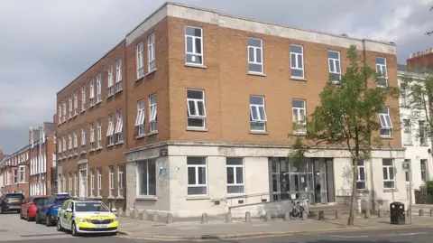 BBC The corner view of a building - it is red brick on top and white brick on the bottom. A police car is at the side of the building.