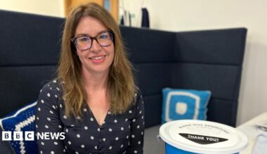 Rachel is sat on a high-backed navy chair in an office, surrounded by Diabetes UK promotional material. She is wearing a navy, floral blouse. She has long brown hair and is wearing dark-rimmed glasses and is smiling at the camera.