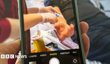A photograph of a phone filming a patient in hospital. In the viewfinder of the phone you can see a patient's arm and a nurse giving him a drug and performing a medical procedure.
