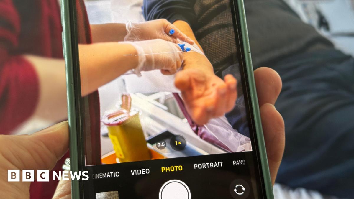 A photograph of a phone filming a patient in hospital. In the viewfinder of the phone you can see a patient's arm and a nurse giving him a drug and performing a medical procedure.