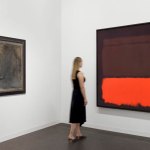A woman looks at an abstract color field painting by Rothko at right. At left is a muddied painted portrait of a man by Giacometti.