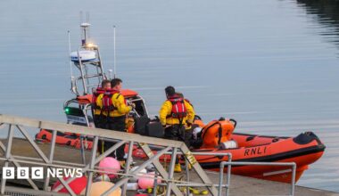 A still harbour with an orange 'RNLI' boat and four men in yellow jackets and red life jackets standing by.