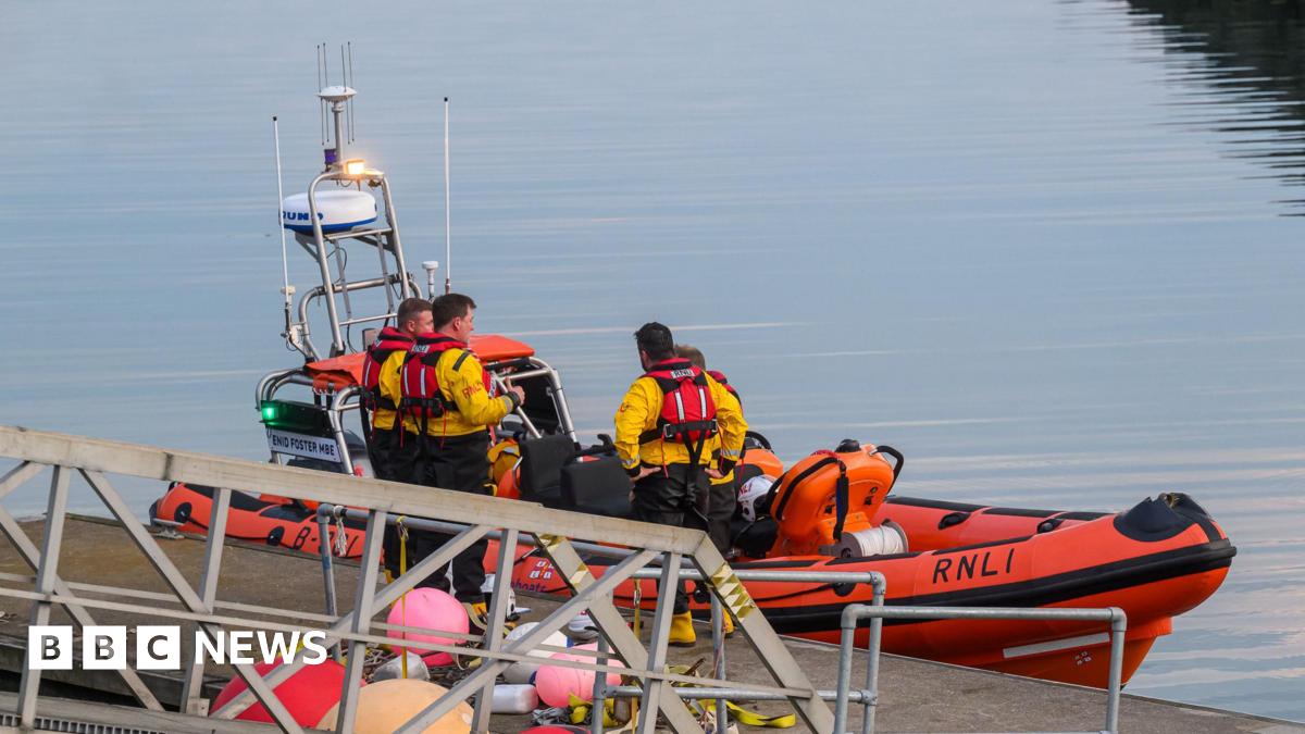 A still harbour with an orange 'RNLI' boat and four men in yellow jackets and red life jackets standing by.