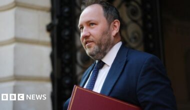 Ian Murray with dark hair and a greying beard walks in front of a stone building. He is wearing a blue suit and tie with a white shirt, and carrying a red folder.
