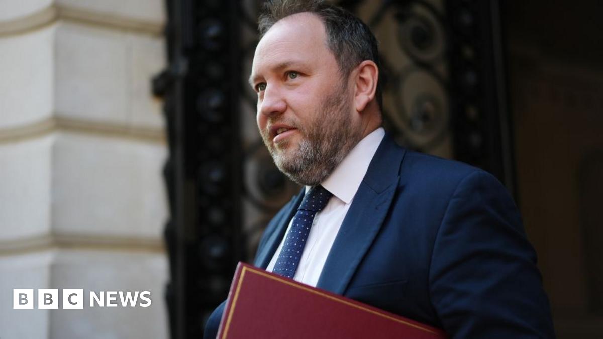 Ian Murray with dark hair and a greying beard walks in front of a stone building. He is wearing a blue suit and tie with a white shirt, and carrying a red folder.