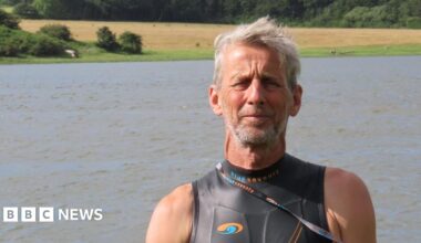Ric Cooper of The Cleddau Project ready to go for a swim in the Western Cleddau river.  He's standing in front of the river wearing a black, sleeveless wetsuit. He has grey short hair and grey stubble.