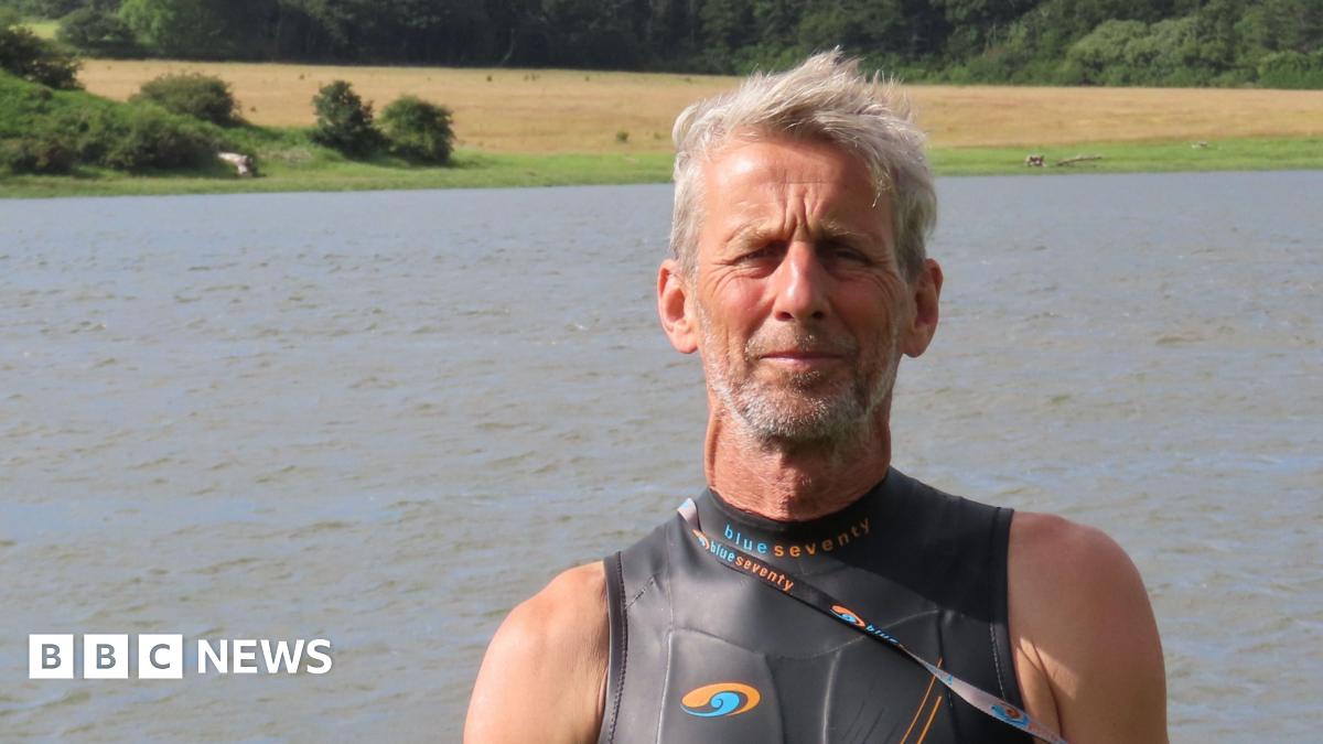 Ric Cooper of The Cleddau Project ready to go for a swim in the Western Cleddau river.  He's standing in front of the river wearing a black, sleeveless wetsuit. He has grey short hair and grey stubble.