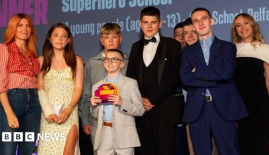 A group of nine people on a brightly lit stage. They are all dressed smartly and looking at the camera. The young boy in the middle of the group is wearing a grey suit and holding an award in his hands.