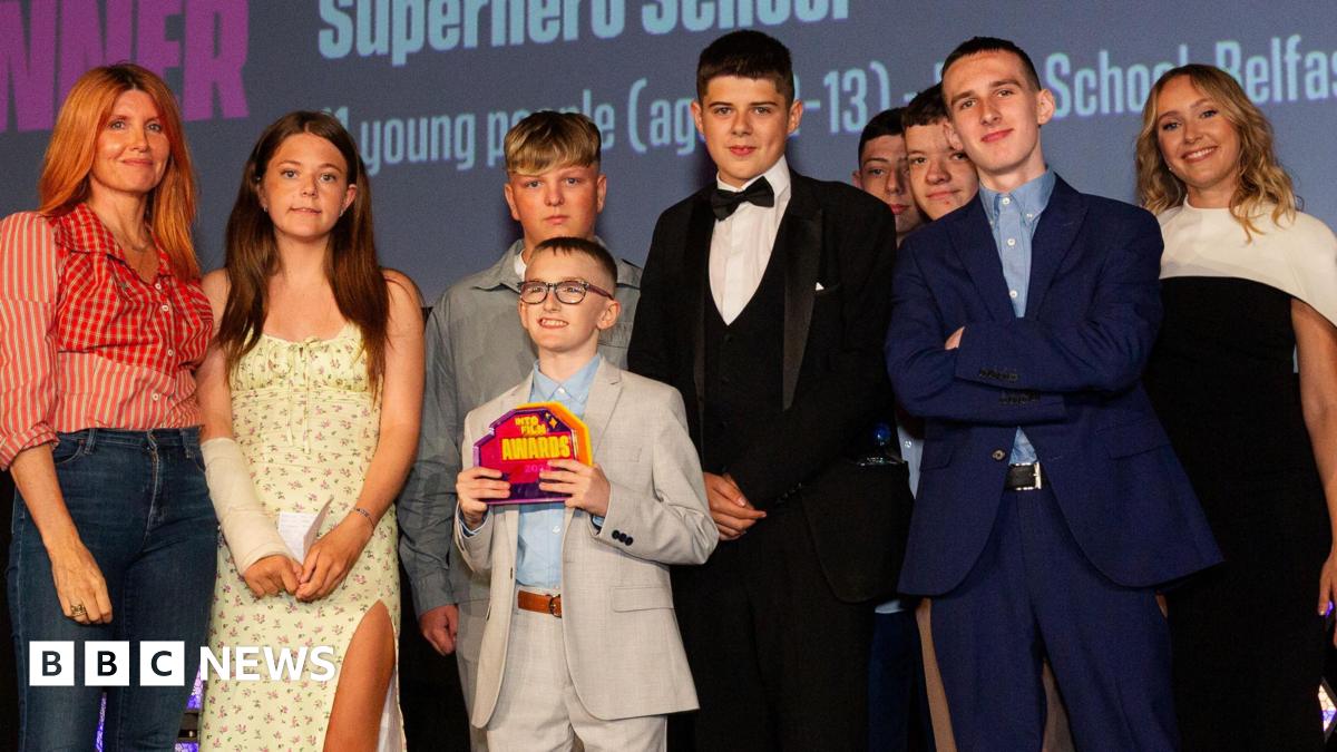 A group of nine people on a brightly lit stage. They are all dressed smartly and looking at the camera. The young boy in the middle of the group is wearing a grey suit and holding an award in his hands.