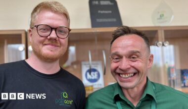 Two men stand side by side, both smiling. On the left, Aran has ginger hair and a ginger beard. He is also wearing a pair of rounded burgundy glasses. Grant, an older man, has brown hair and wears a green shirt. Behind them, a blurred view of a cabinet with a display of company awards.