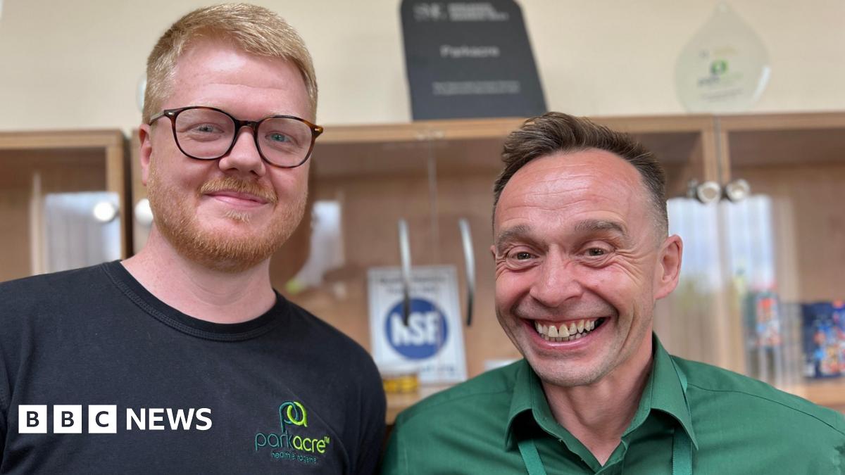Two men stand side by side, both smiling. On the left, Aran has ginger hair and a ginger beard. He is also wearing a pair of rounded burgundy glasses. Grant, an older man, has brown hair and wears a green shirt. Behind them, a blurred view of a cabinet with a display of company awards.