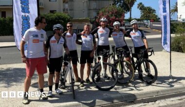 Seven men who have finished a cycling challenge from Leeds to Nice, they are standing a row with their arms around each other, some with their bikes, some wearing helmets, all smiling.