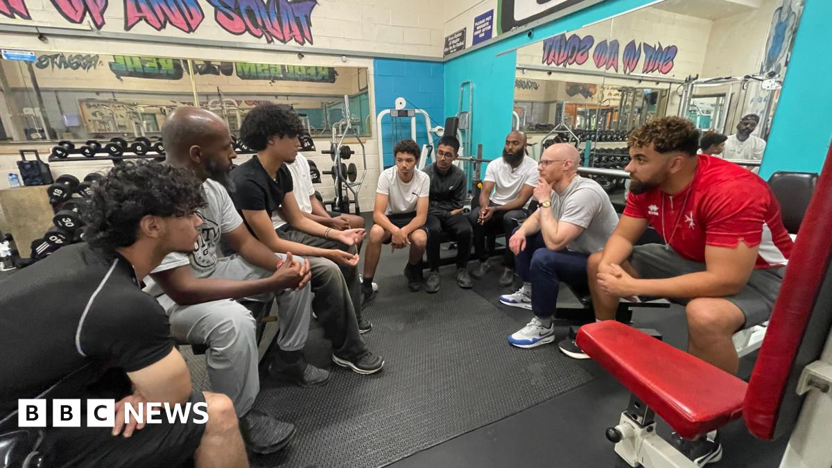 A group of young men sit in a circle in a gym listening to one-another