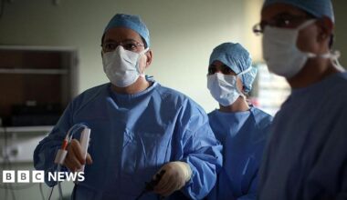 A surgeon and his theatre team perform key hole surgery to remove a gallbladder at the Queen Elizabeth hospital in Birmingham. The doctors are all wearing blue overalls and white masks and staring off camera. The main surgeon is holding a medical device.