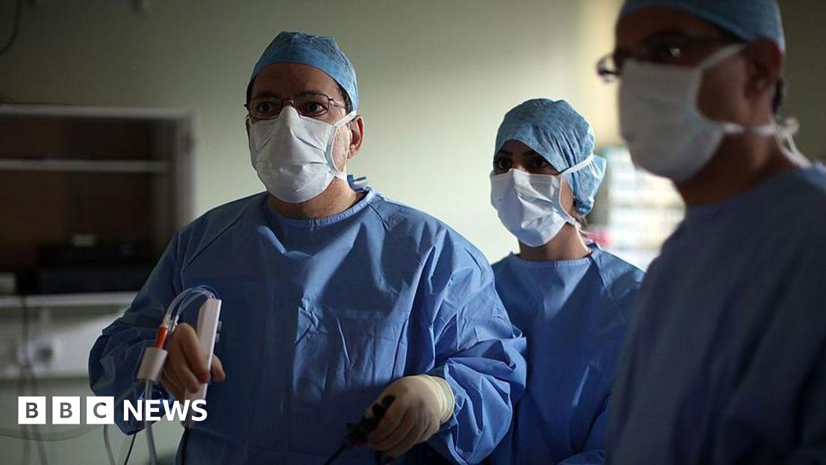 A surgeon and his theatre team perform key hole surgery to remove a gallbladder at the Queen Elizabeth hospital in Birmingham. The doctors are all wearing blue overalls and white masks and staring off camera. The main surgeon is holding a medical device.
