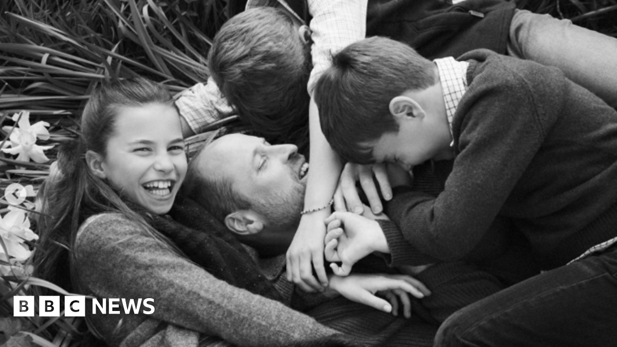 A black and white photo of Prince William lying on the grass - his three children hug him while Charlotte looks at the camera and smiles