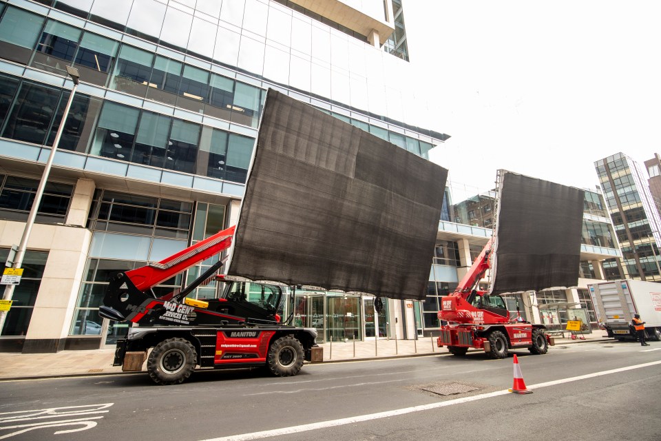 Two cranes lifting large black screens in front of a building.