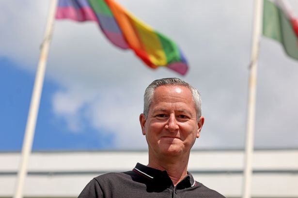 The image shows Mark posing in front of a rainbow pride flag