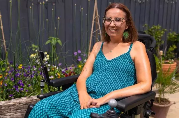 Emma Bray photographed in her wheelchair wearing a blue dress outside in a garden with flower beds