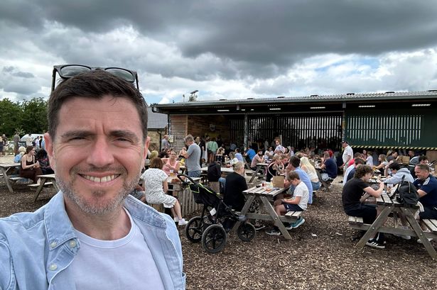 Steffan Rhys poses for a selfie in front of crowds of people sitting at tables outside Jeremy Clarkson's Diddly Squat Farm shop