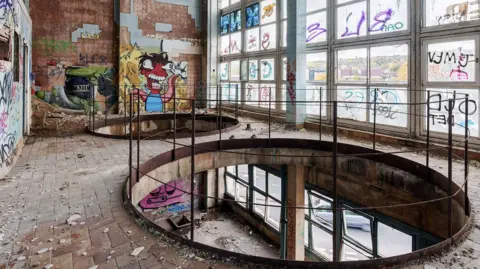 Capital&Centric Inside a derelict building. A full-wall glass window is covered in graffiti on the inside and outside. There are tiles falling off the walls and dust from the degrading building. Mostly graffiti tags but on the far wall is a painting of a red octopus type monster. 