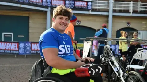 Dave Eastham Dave Eastham is sitting in his wheelchair wearing a sports kit and trainers. He has brown hair and is wearing a blue sports top, smiling at the camera