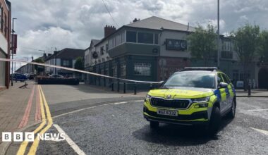 A marked police car is in the foreground. White police tape is across the street. In the background you can see buildings on either side of the street and double yellow lines along the left hand side as far as the eye can see.