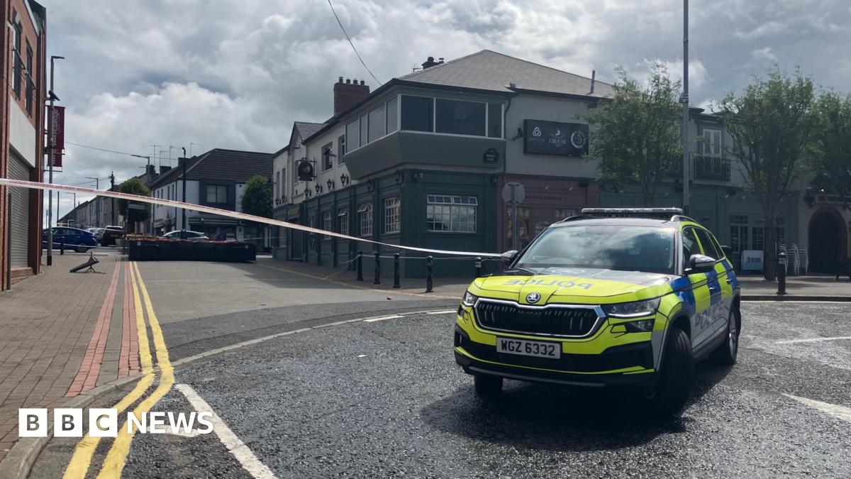 A marked police car is in the foreground. White police tape is across the street. In the background you can see buildings on either side of the street and double yellow lines along the left hand side as far as the eye can see.